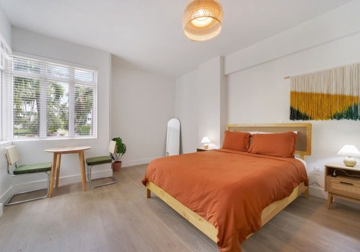 Bright bedroom with orange bedspread, white walls, desk by window, and abstract wall art.