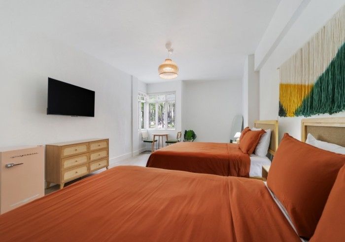 Bright bedroom with two orange-covered beds, wall-mounted TV, dresser, and green-yellow wall art