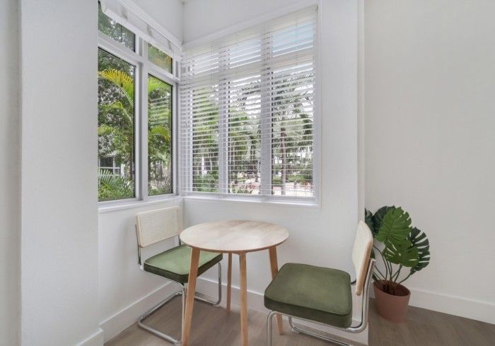 Bright corner nook with white blinds, round wooden table, green cushions, and a potted plant.