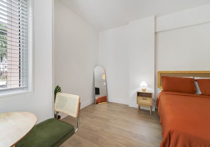 Bright minimalist bedroom with orange bedspread, mirror, window, and small chair by the wall