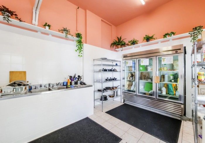 Small bakery or café interior with white counter, display fridge, and potted plants on a peach wall