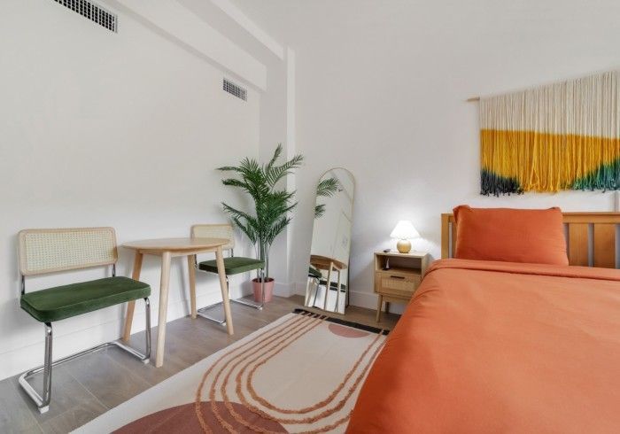 Bright bedroom with an orange bed, green chair, small table, plant, mirror, and striped rug