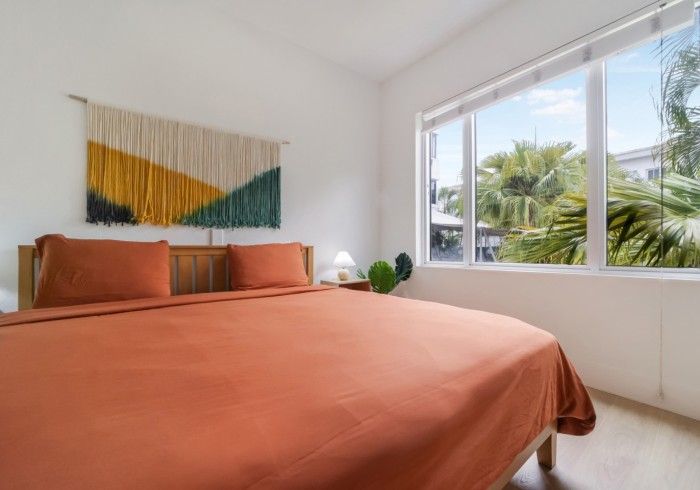 Bright bedroom with orange bedspread, abstract wall art, and large sunlit window overlooking palm trees