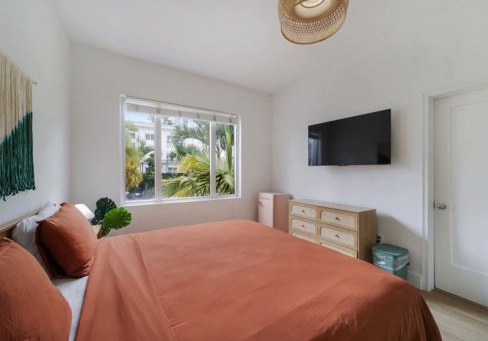 Bright bedroom with coral bedspread, wall-mounted TV, window view, and white door on the right