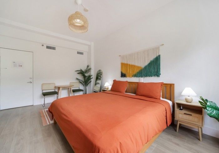 Bright bedroom with orange bedspread, wooden nightstands, desk, plant, and abstract wall art
