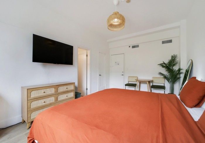 Bright bedroom with orange bedspread, wall-mounted TV, dresser, and potted plant