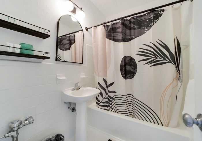 Small white bathroom with pedestal sink, mirror, tub, and black-and-white patterned shower curtain