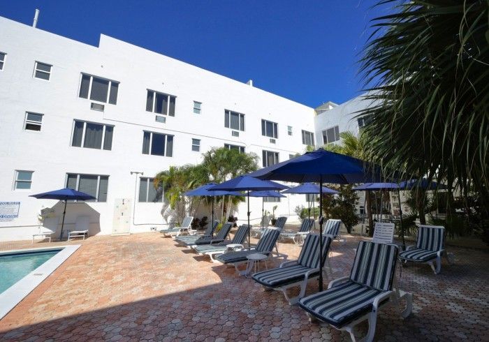 Poolside lounge chairs and blue umbrellas beside a white hotel building under a clear sky