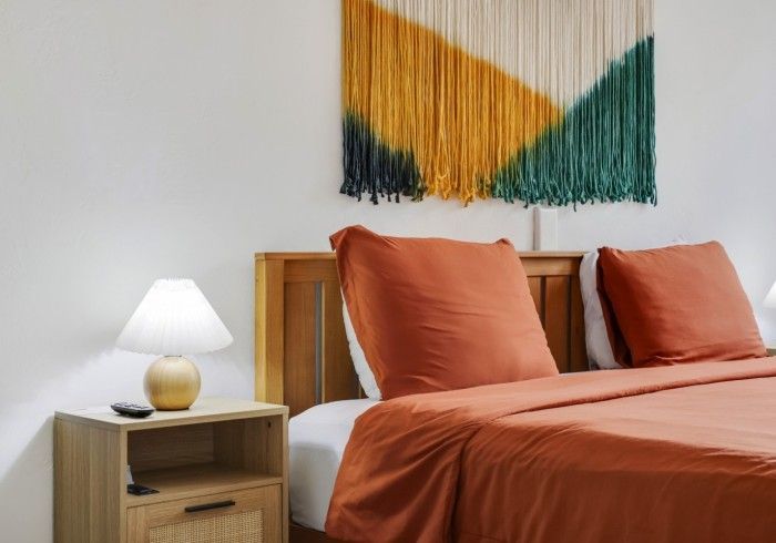 Modern bedroom with rust-orange bedding, wooden nightstand, white lamp, and abstract wall hanging.