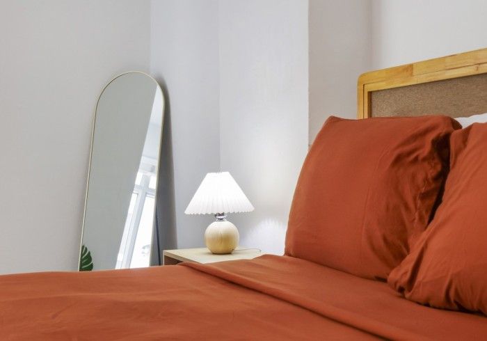 Orange bed with pillows beside a small white lamp and leaning mirror in a bright bedroom