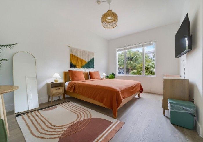 Bright bedroom with orange bedspread, rug, nightstand, wall art, and a window with trees outside