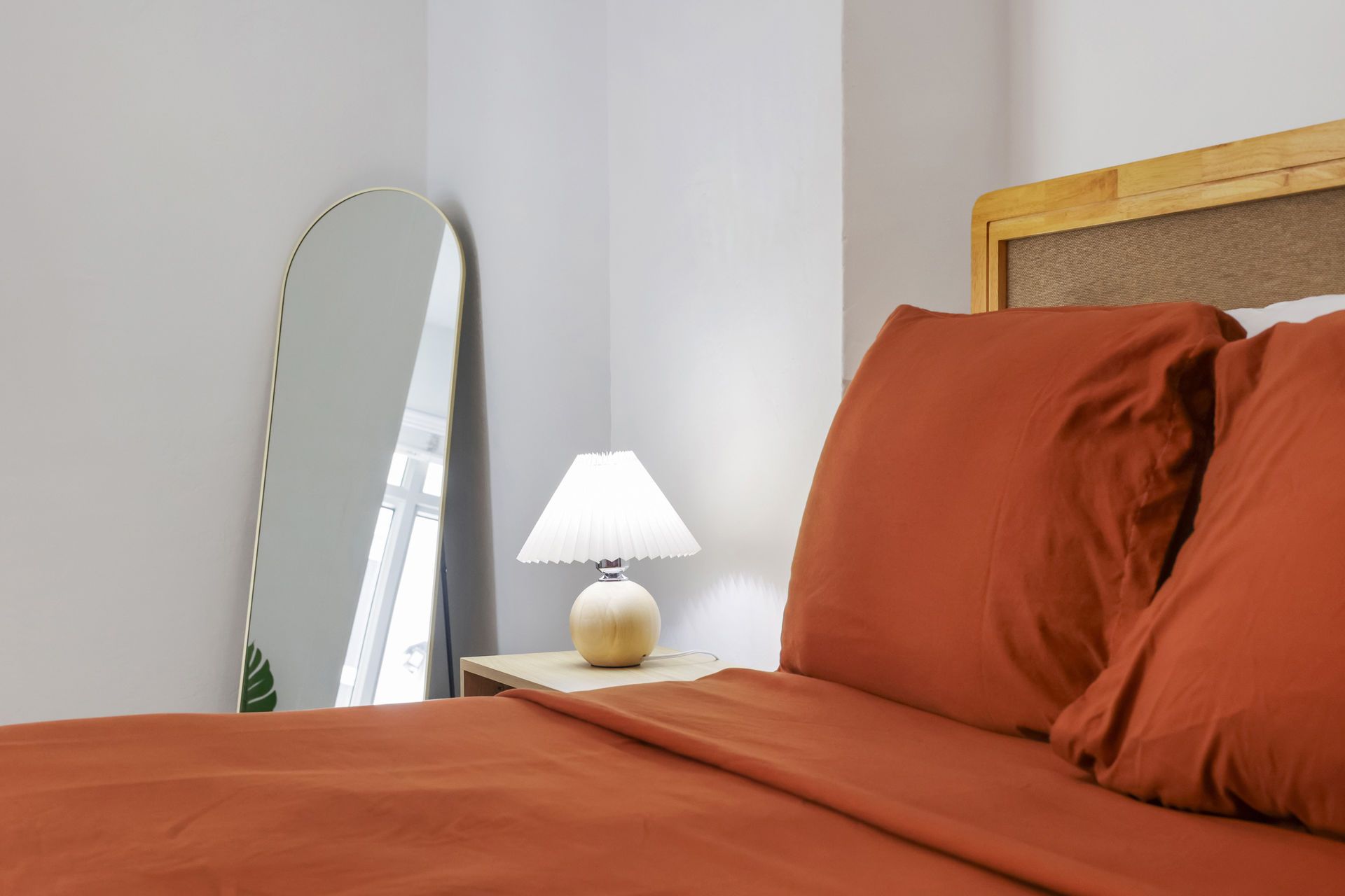 Orange bed with pillows beside a small white lamp and a leaning mirror in a bright bedroom