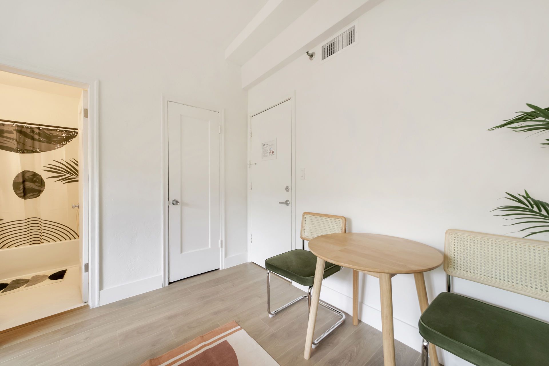Bright minimalist room with a small round table, green chairs, white walls, and an open doorway to a patterned bathroom