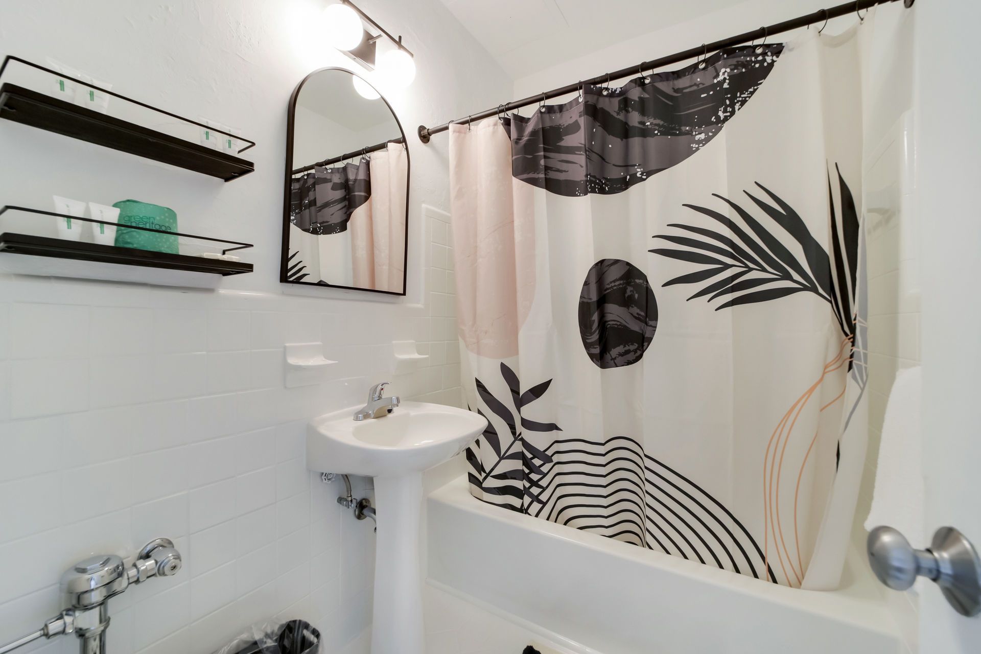 Small bathroom with shower curtain, wall mirror, sink, shelves, and white tiled walls.