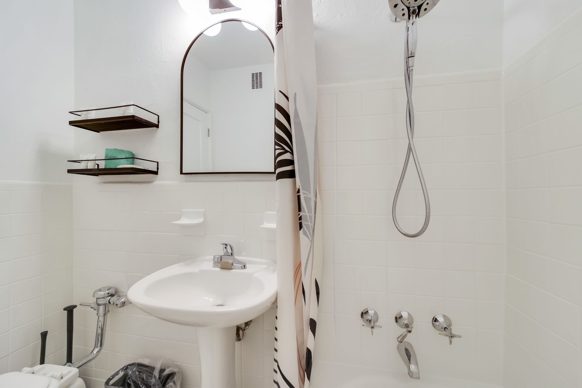 Small white bathroom with sink, shelves, mirror, and shower curtain in a tiled shower area