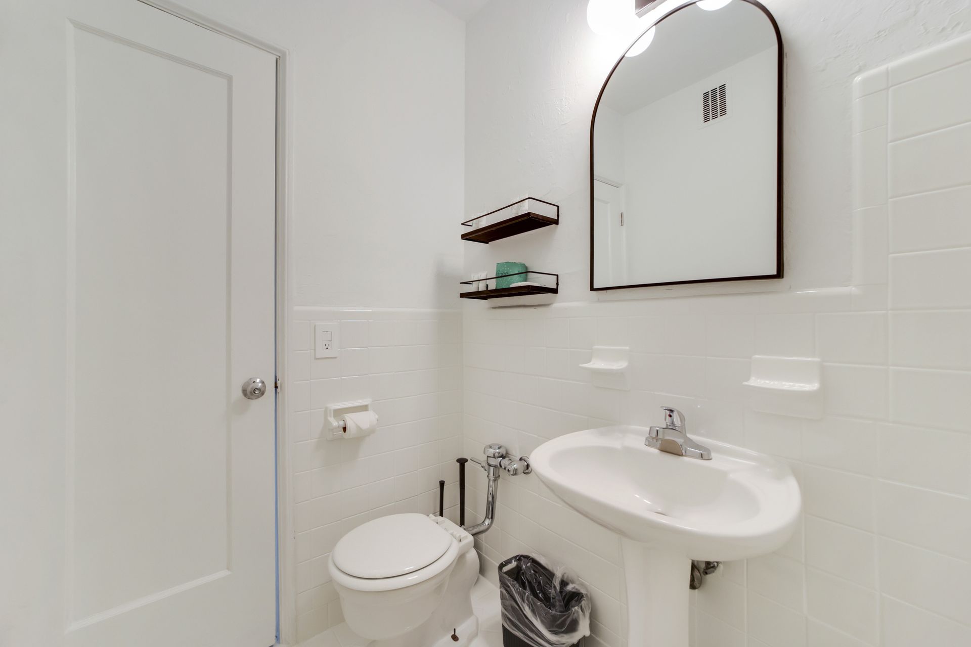 Small white bathroom with toilet, pedestal sink, mirror, and wall shelves.