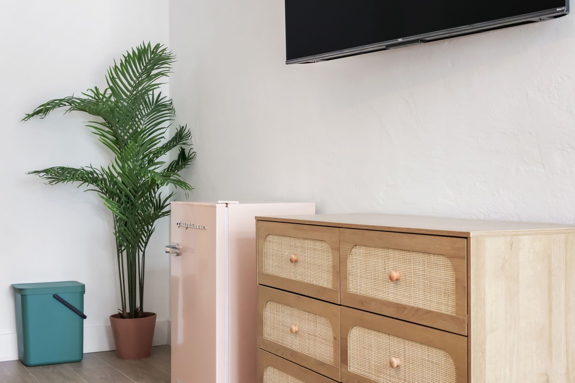 A potted palm and blue bin beside a light wood cabinet in a bright room with a wall-mounted TV.