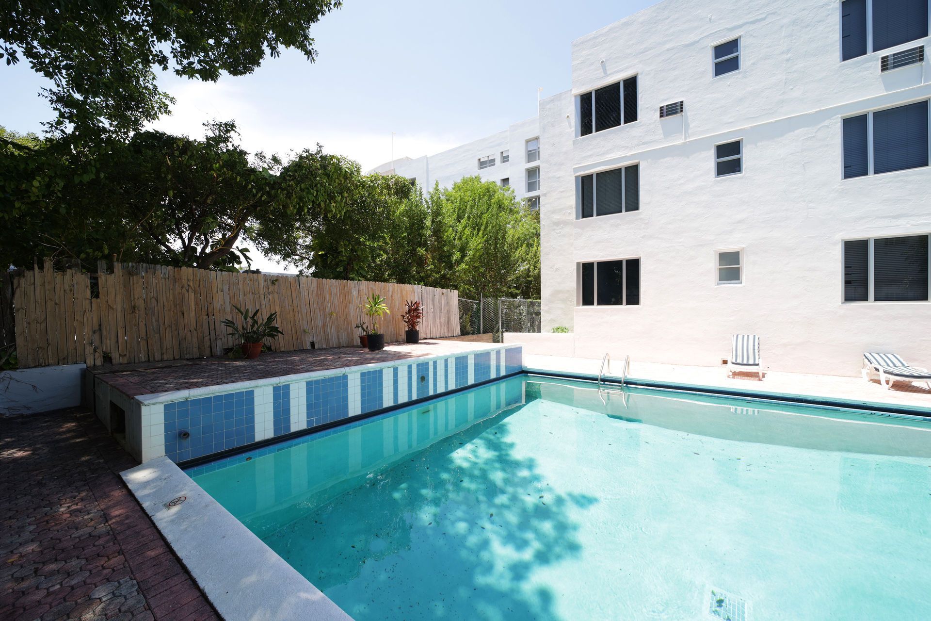 Outdoor swimming pool beside a white apartment building, with blue water and trees along the fence