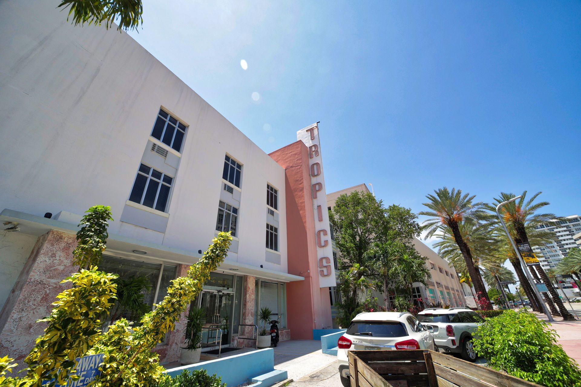 Sunlit hotel entrance with pink vertical sign, palm trees, and parked cars