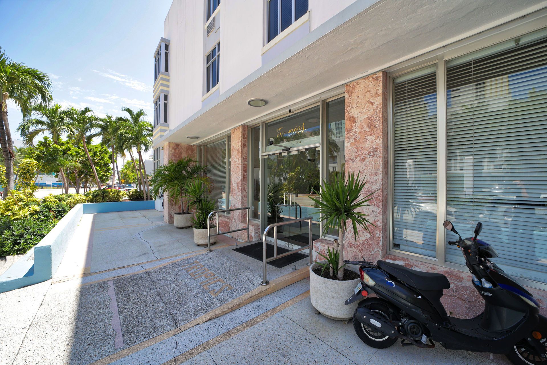 Modern storefront with glass windows, potted plants, outdoor seating, and a parked scooter along a sunny walkway