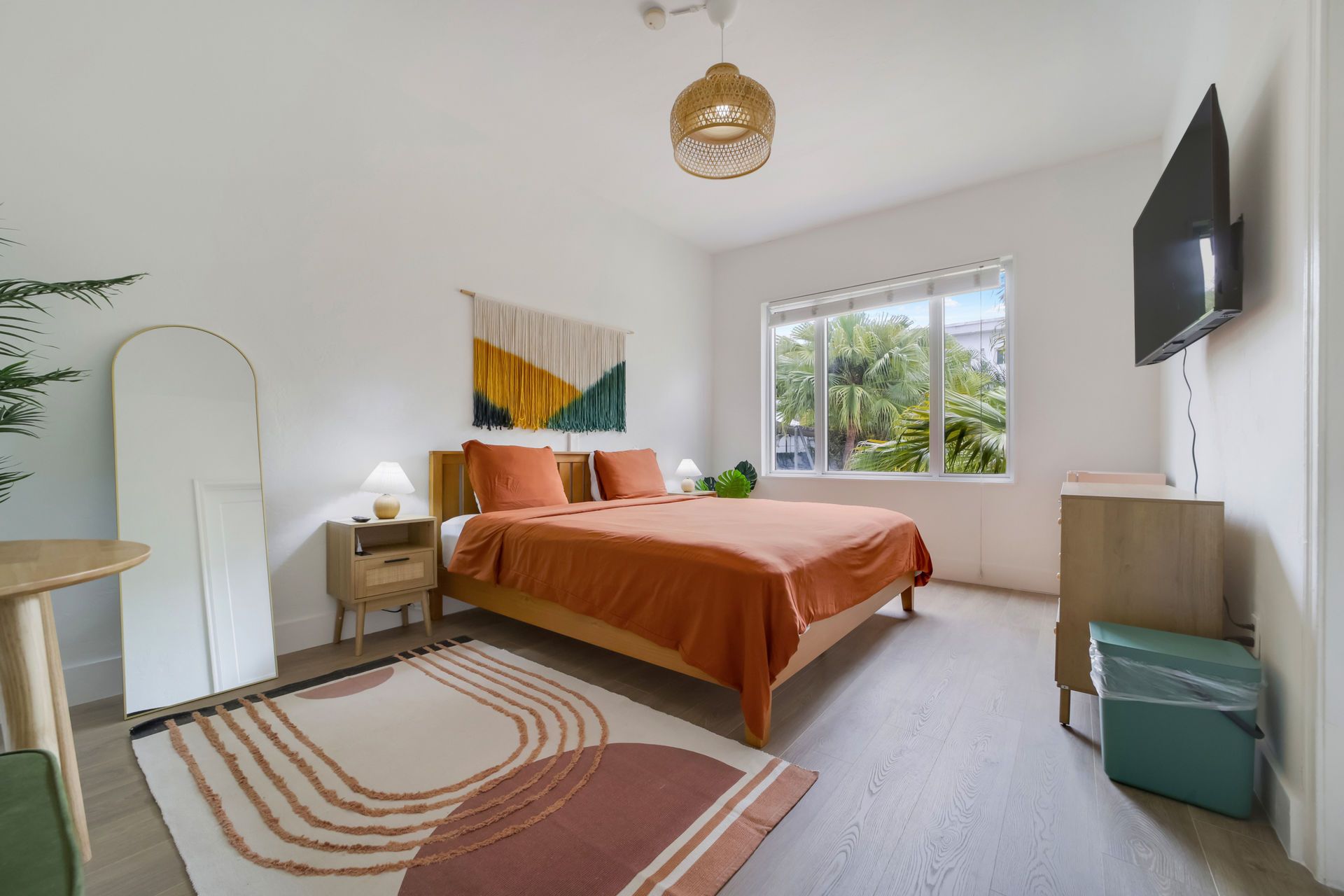 Bright bedroom with orange bedspread, window, wall-mounted TV, and rainbow rug
