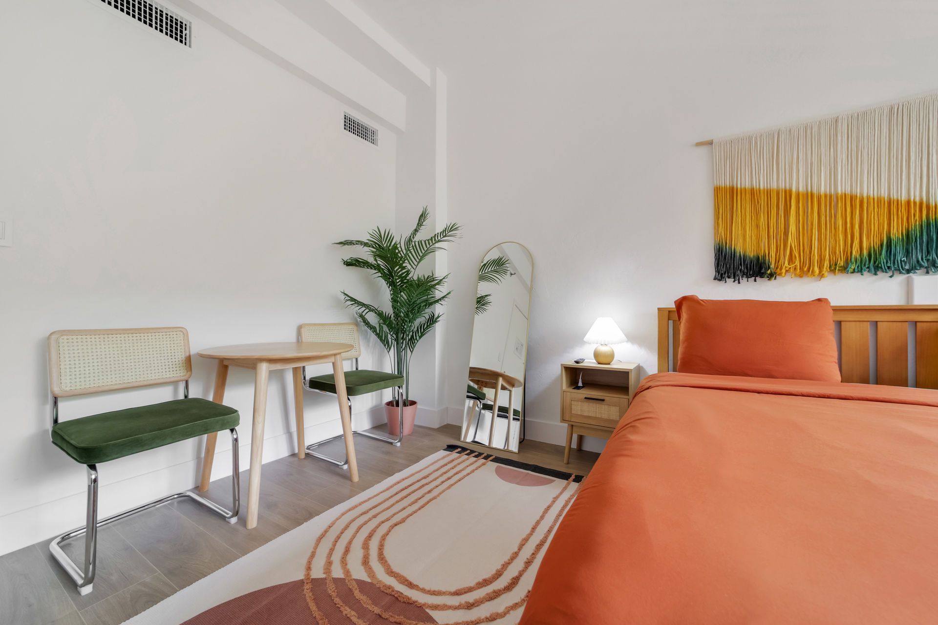 Minimal bedroom with orange bed, small desk, potted plant, and striped rug in a bright white room