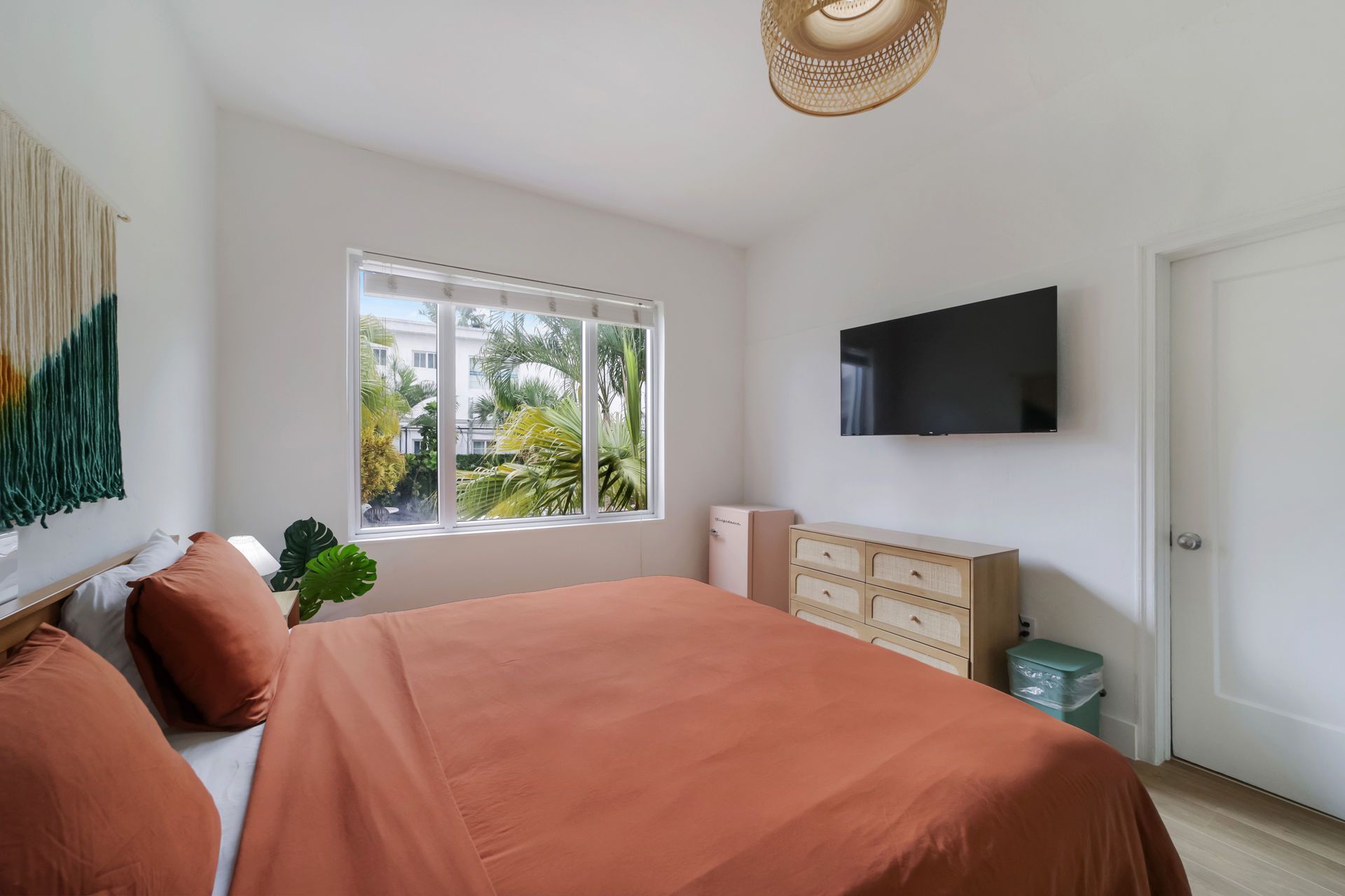 Minimal bedroom with coral bedspread, wall-mounted TV, dresser, window, and woven pendant light.