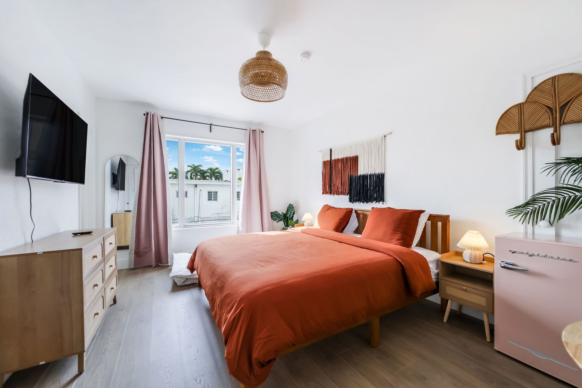 Bright bedroom with orange bedspread, pink curtains, wall TV, and balcony doors letting in daylight