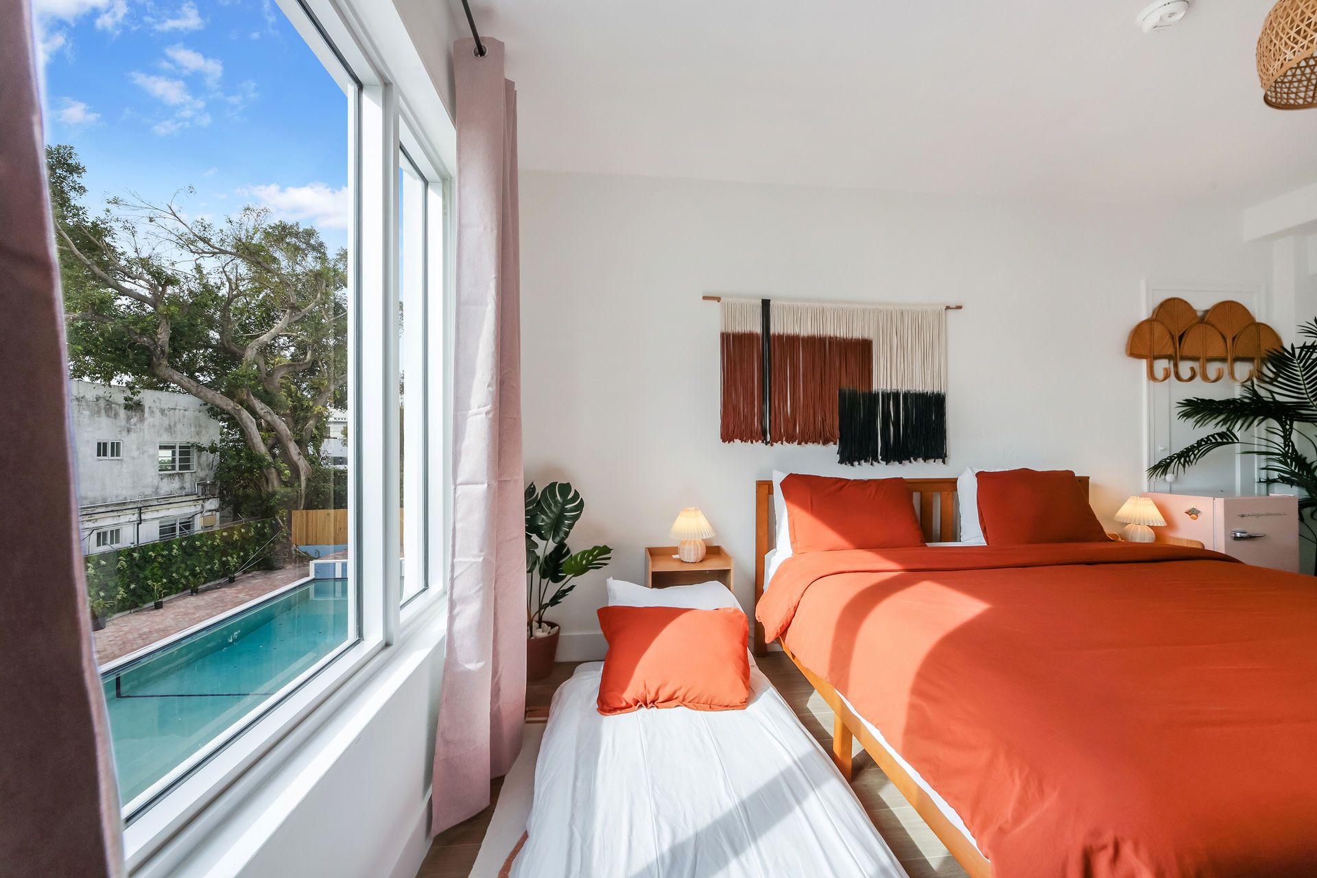 Bright bedroom with orange bedding, pink curtains, and a large window overlooking a pool and trees