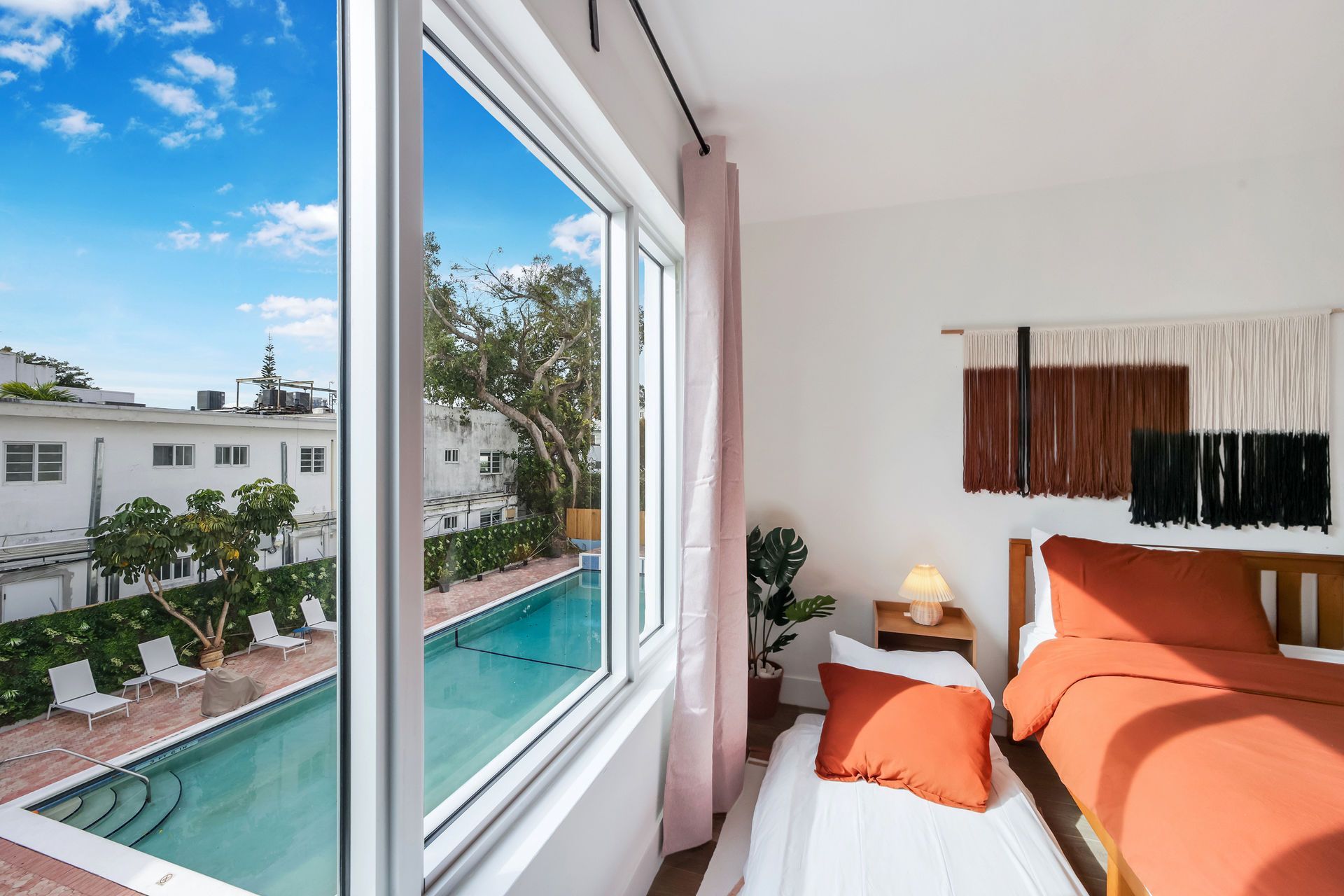 Bright bedroom with orange bedding, large window, and a view of an outdoor pool and white buildings