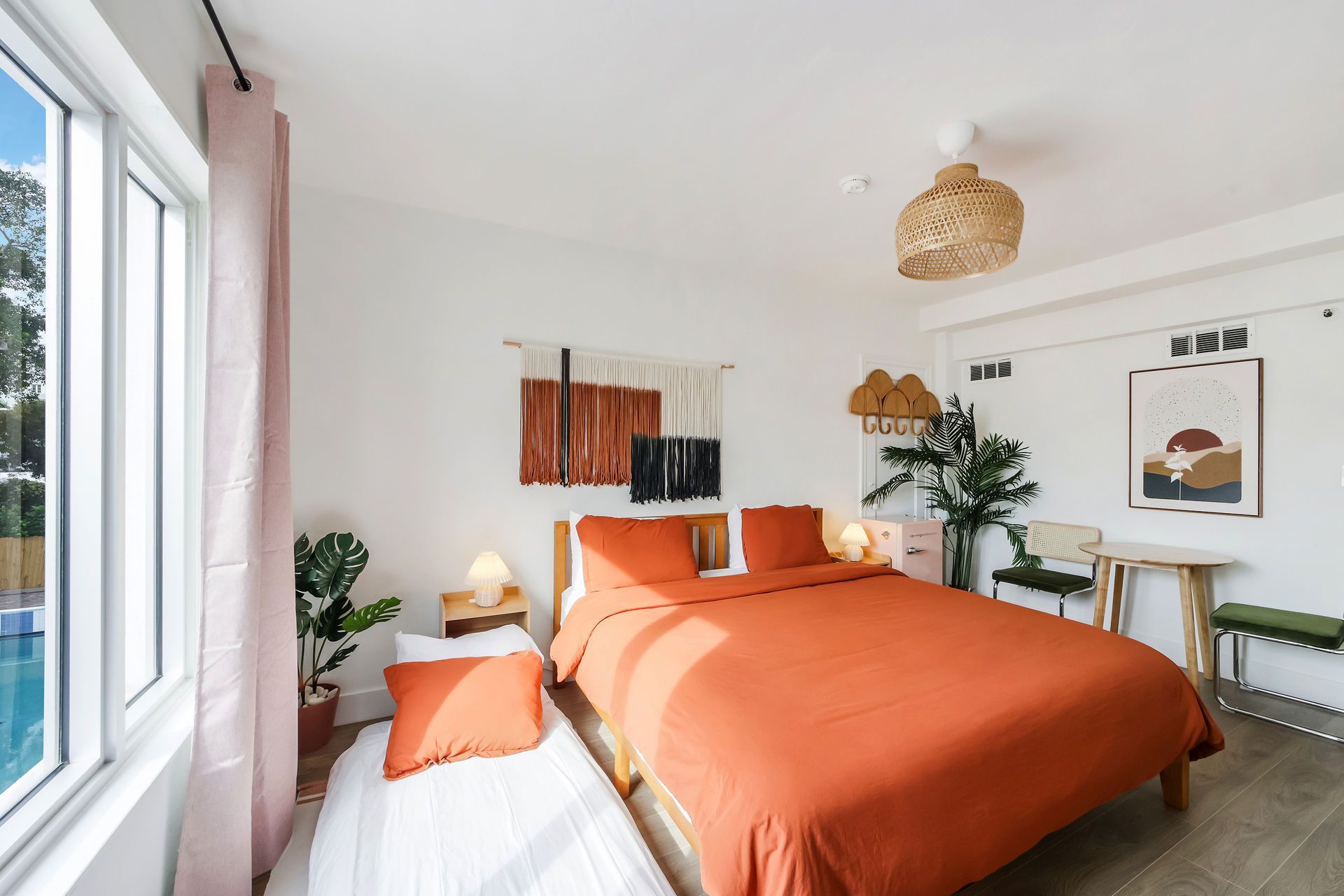 Bright bedroom with orange bedding, large window, and plants in a modern white interior.