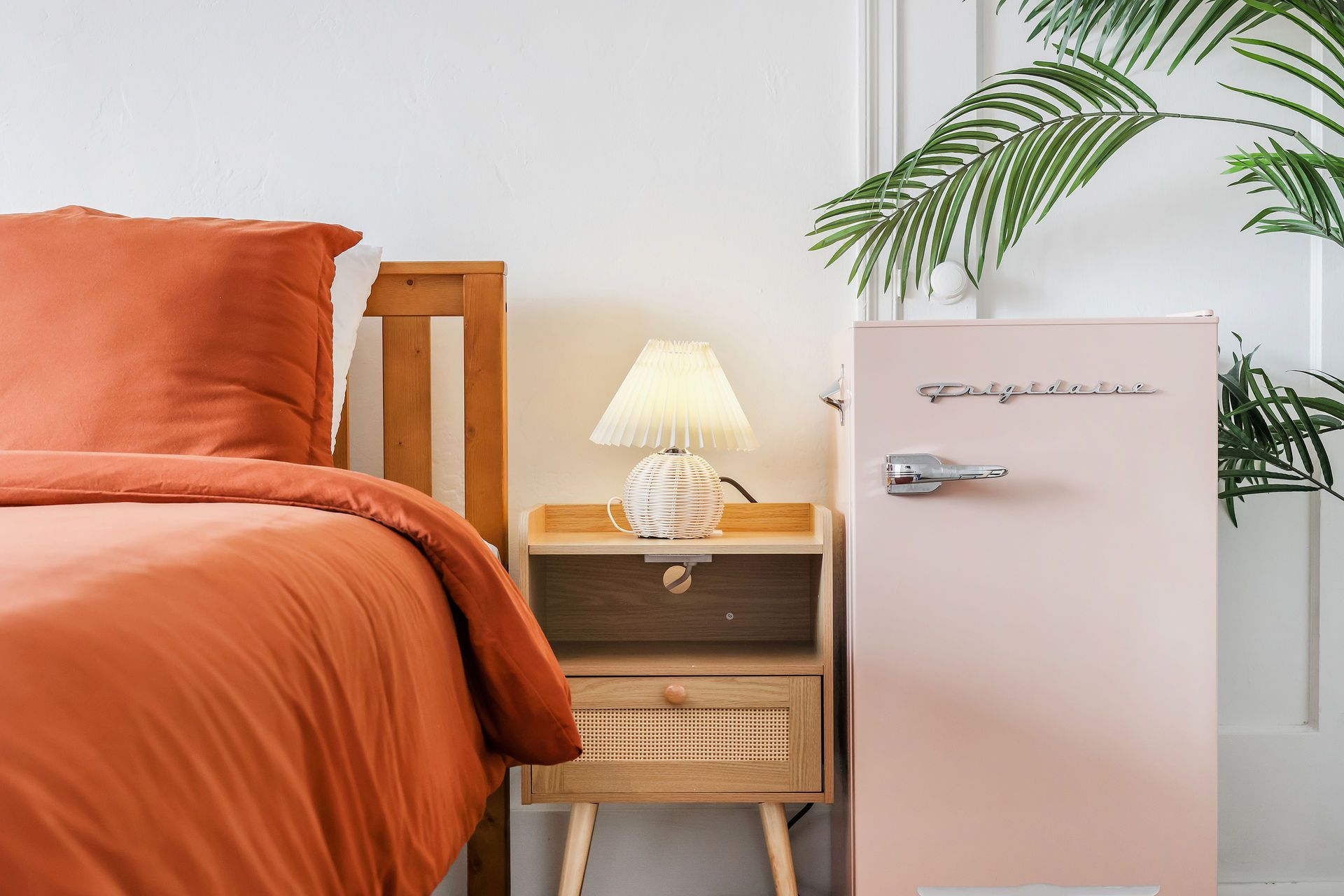 Orange bed beside a pink mini fridge and lit bedside lamp in a bright bedroom