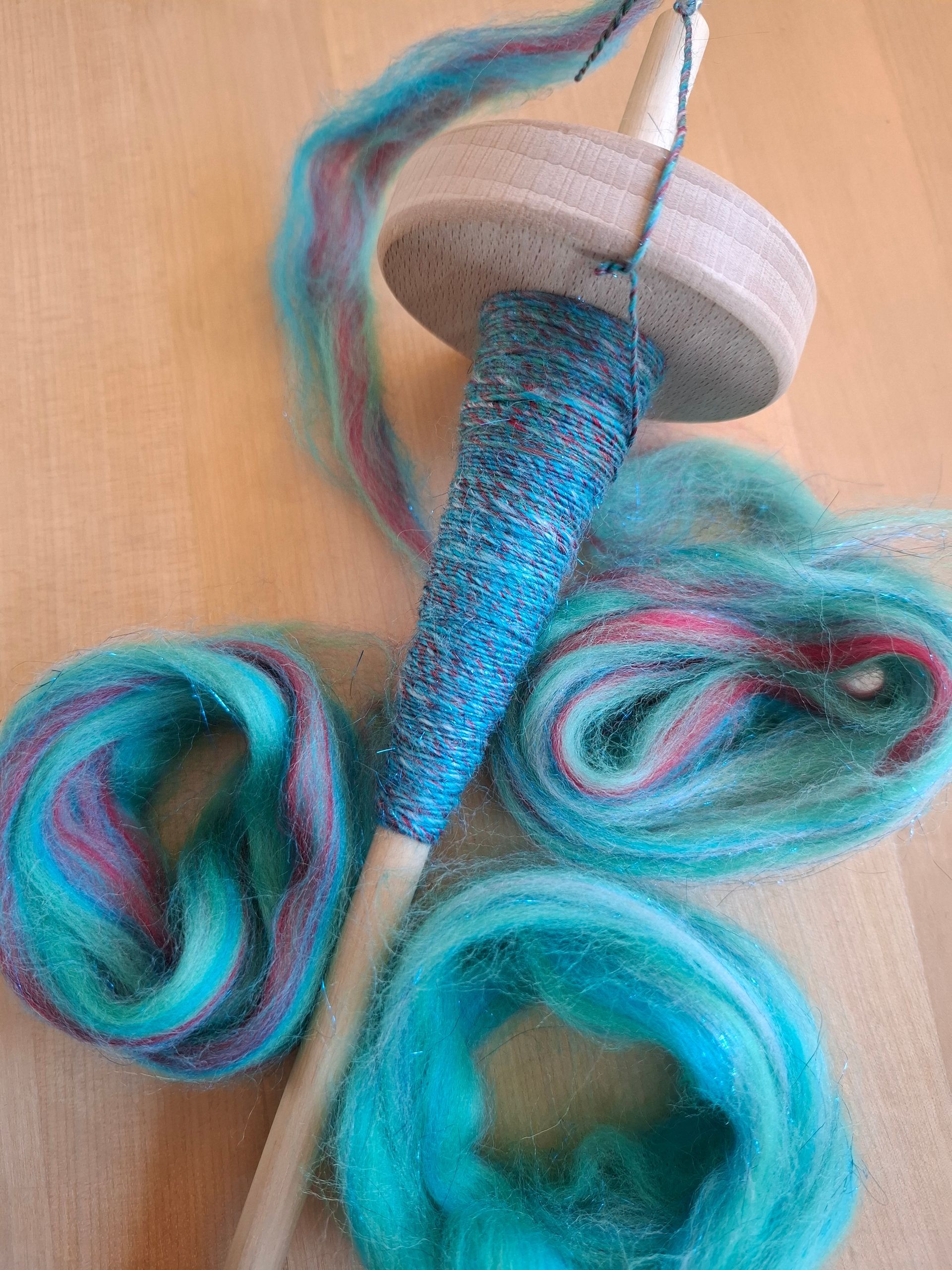 Een handspindel met blauwe vezels eromheen gewikkeld en twee rollen blauwe vezels op een houten oppervlak.