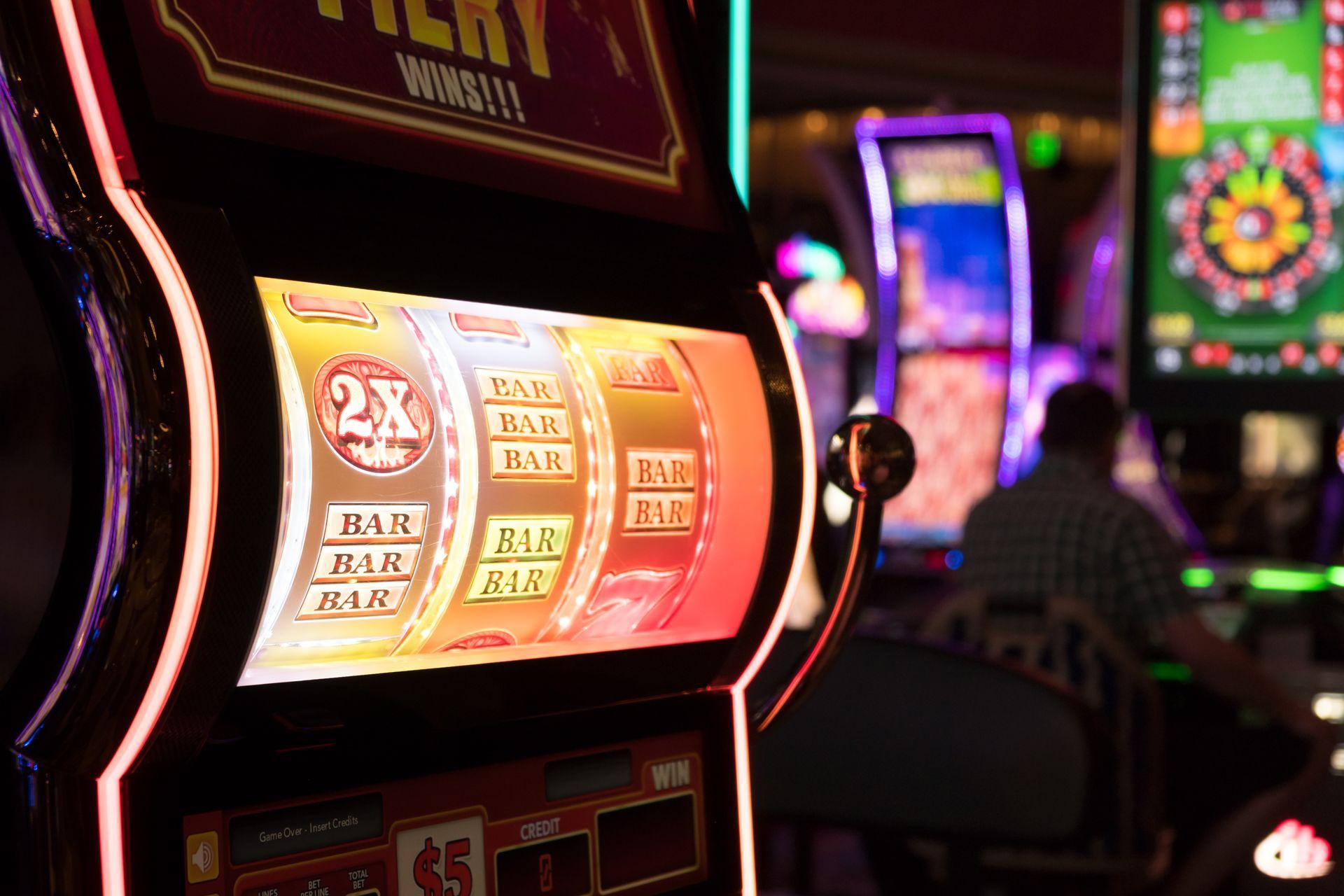 Slot machine glowing in a colorful casino, with blurred machines and a seated player in the background