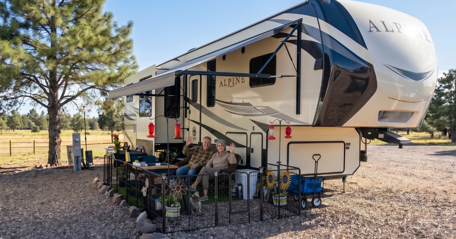 Camper trailer with awning and outdoor chairs in a gravel campsite, trees and open field nearby