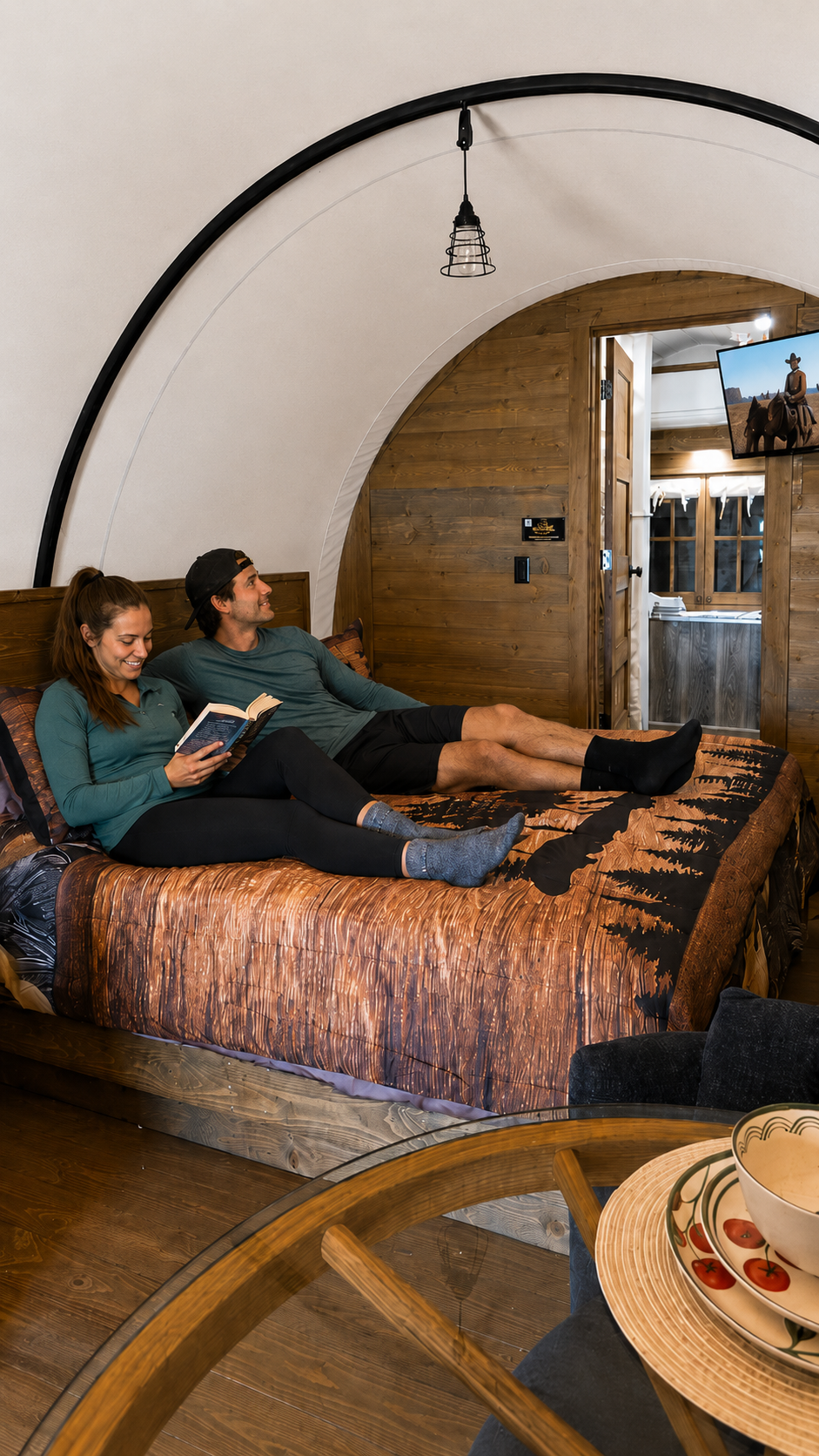 Two people relaxing on a bed inside a cozy wooden pod with an open door and mountain view.