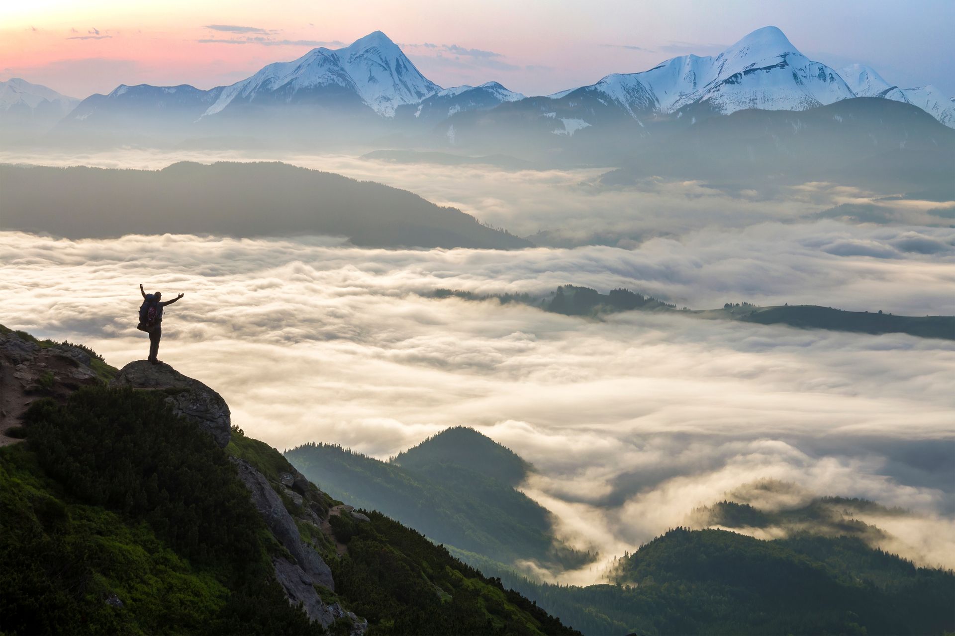 Hiker on a cliff above a sea of clouds with snow-capped mountains in the distance