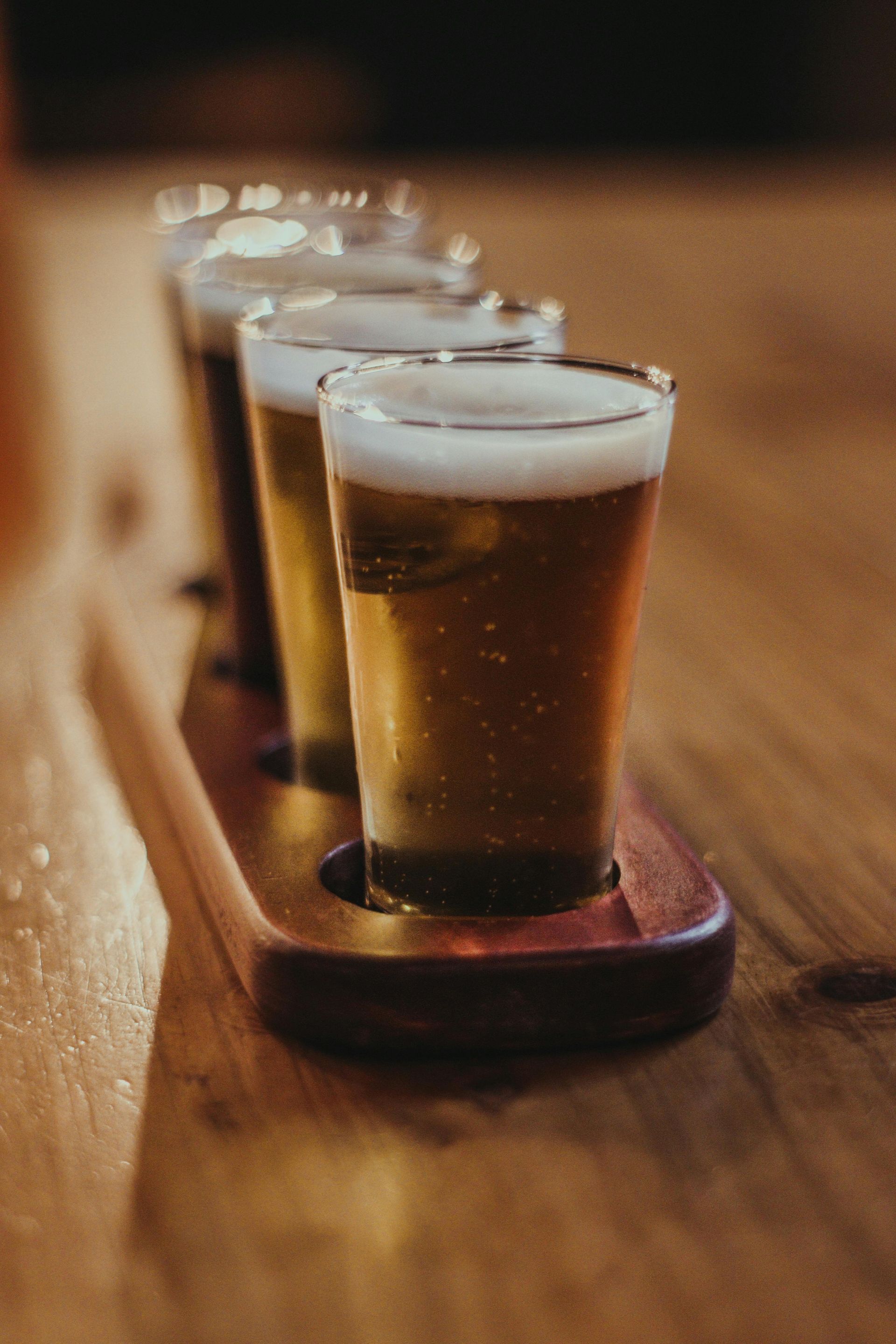 Row of small beer samples on a wooden table, each in a clear tasting glass with foam on top