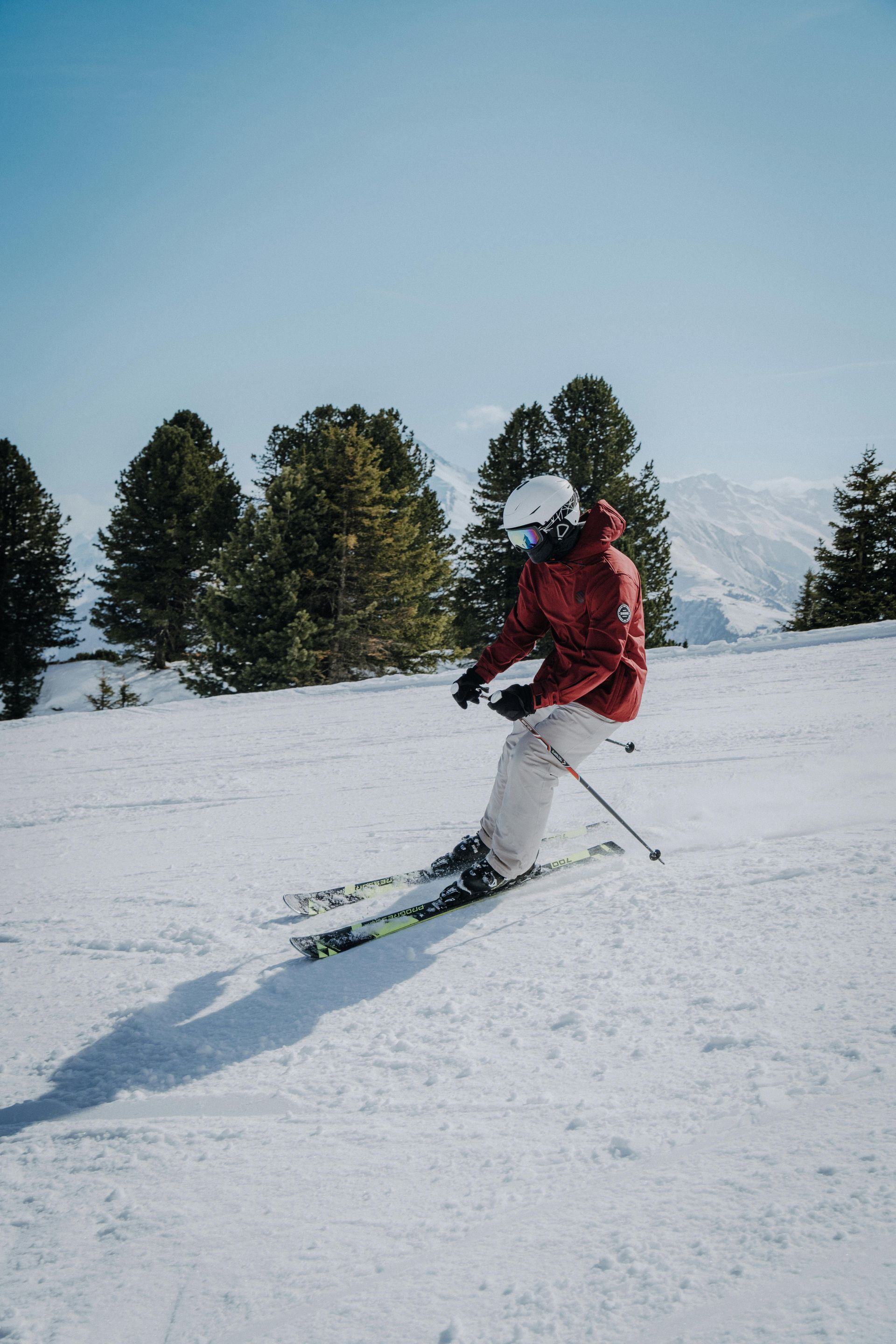 Skier in a red jacket carving downhill on a snowy slope with pine trees and mountains behind.