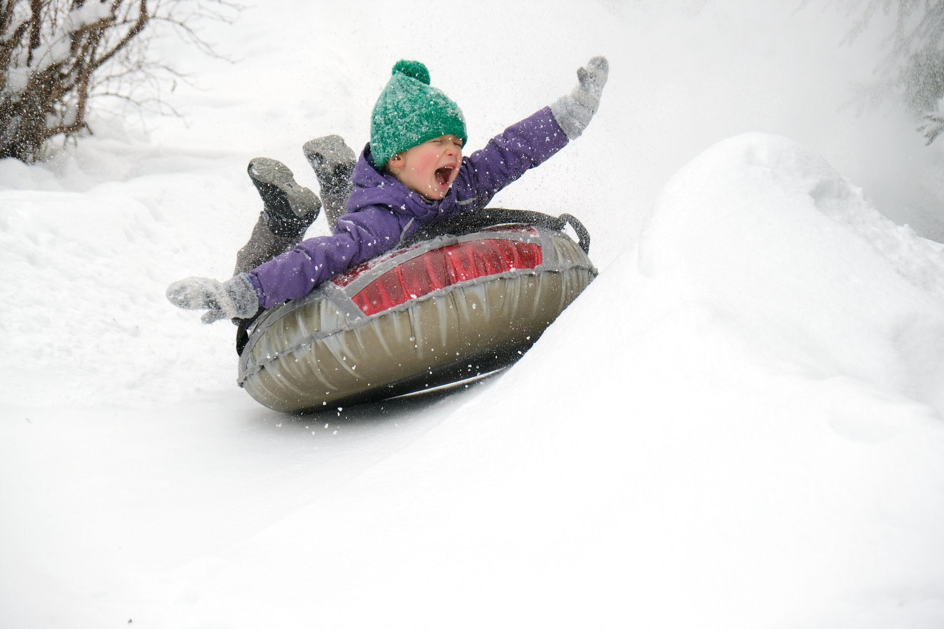 Person sledding on a snowy hill in a gray inflatable tube, wearing a purple jacket and green hat.