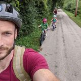 A man wearing a helmet is taking a selfie with three children riding bikes down a road.