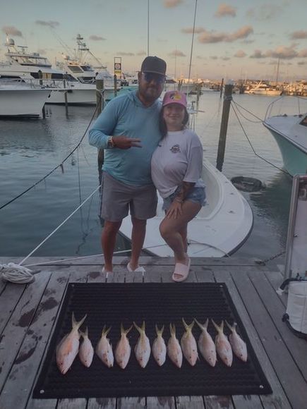 Couple poses with a row of caught fish on a dock, boats in background.
