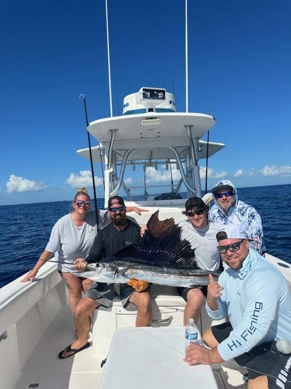 Five people on a boat with a large fish. Clear blue sky and ocean. People smiling, celebrating a catch.