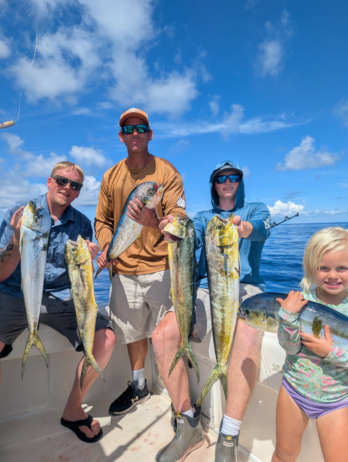 Four people smiling on a boat, holding up freshly caught mahi-mahi fish on a sunny, blue-sky day.