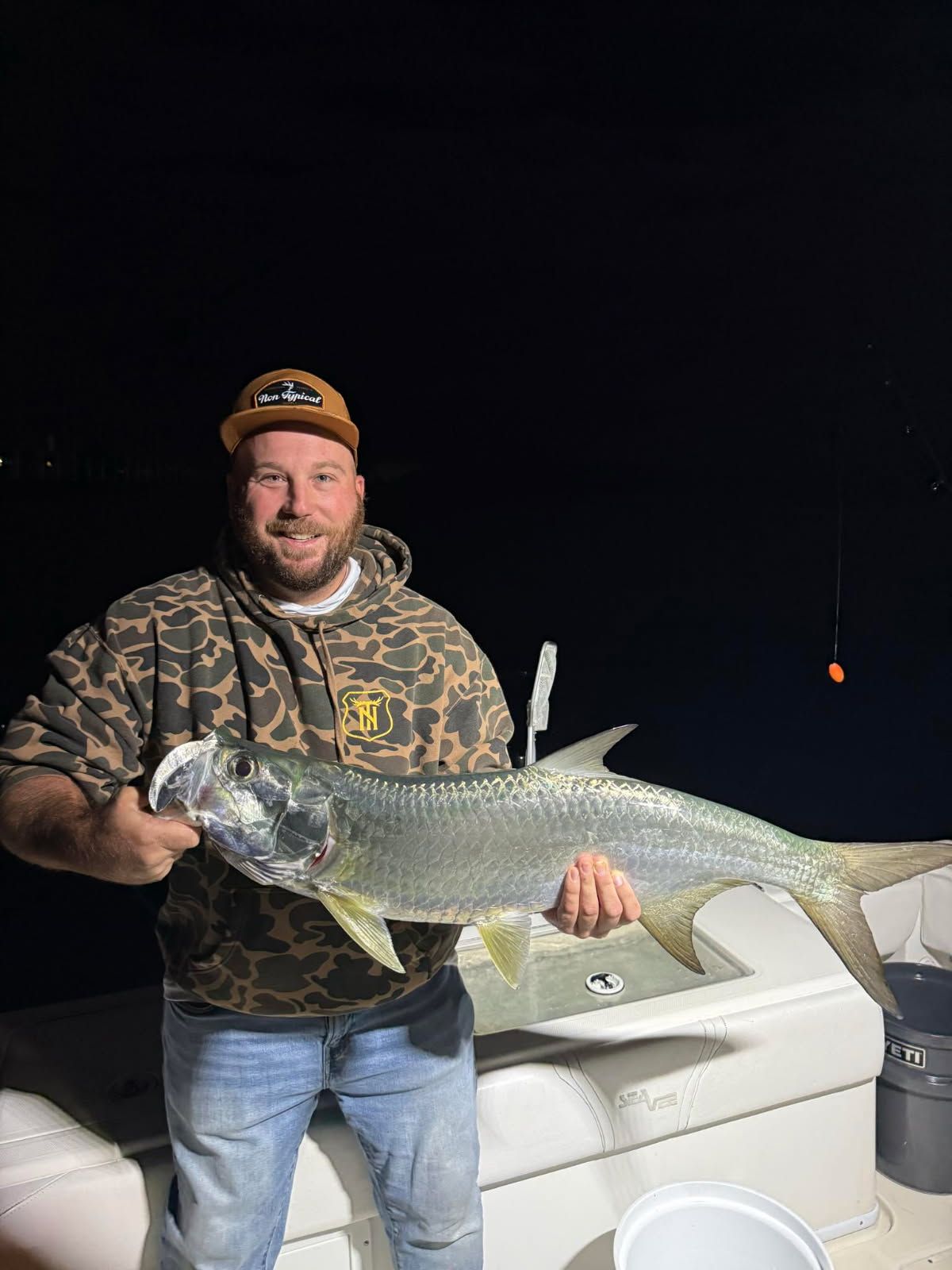 Man on a boat holding a large, silver fish at night. He is wearing a camo hoodie and smiling.