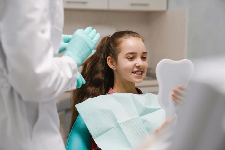 A young girl is sitting in a dental chair looking at her teeth in a mirror.