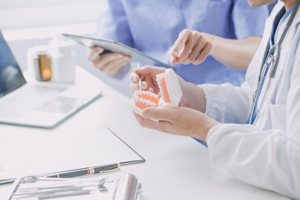 An Oral Hygienist is holding a model of a tooth while a patient looks at a tablet.