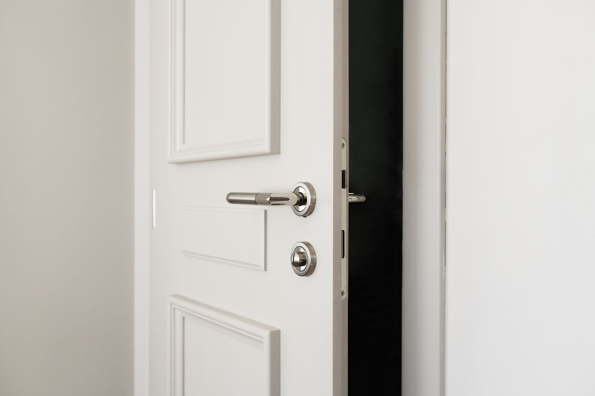Porta bianca socchiusa, ferramenta argentata, apertura su uno spazio buio, ambiente interno.