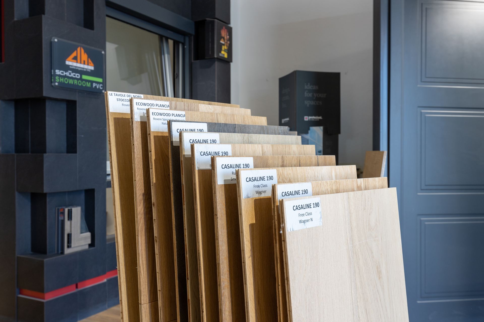 Esposizione di campioni di legno con etichette in uno showroom, accanto a una porta grigio scuro.