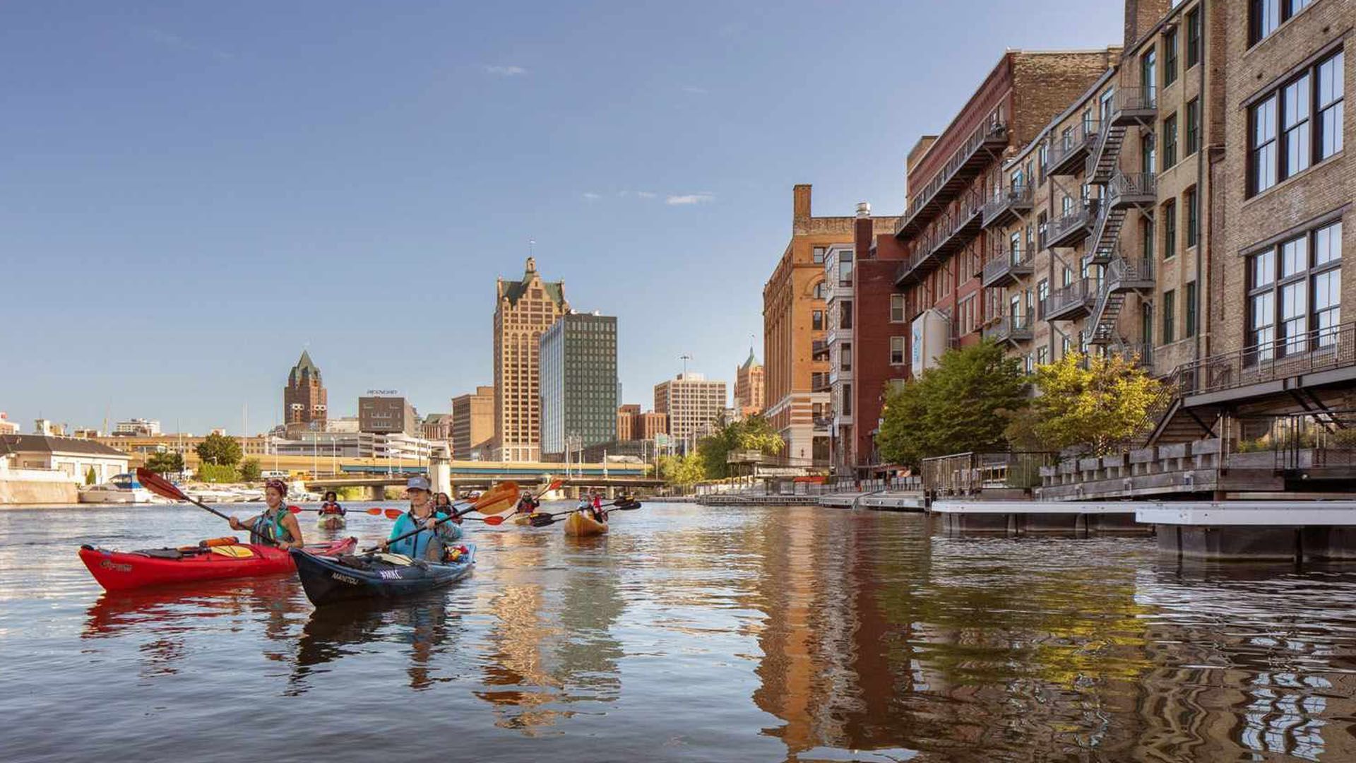 View of Milwaukee RiverWalk with kayakers and boats on a sunny summer day.
